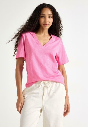 Kvinde med langt krøllet hår, iført en knaldrød V-hals T-shirt og off-white bukser med snøring, stående mod en ensfarvet baggrund.