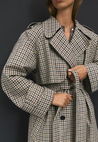 Karierter Trenchcoat in Beige, Braun und Blau. Mit weitem Kragen, übergroßen Ärmeln und einem gebundenen Taillengürtel. Mit Knopf- und Taschendetails.