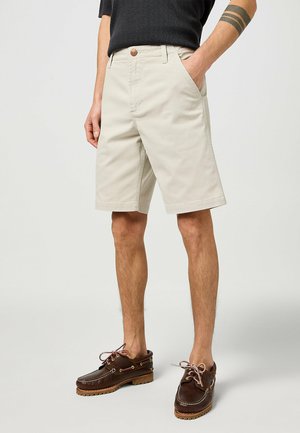 Beige bomullsshorts med två sidofickor, knappstängning och rak skärning, bärs med bruna läderbåtskor.