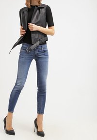 Svart läderväst över en figursydd svart topp, kombinerad med blåa tajta jeans och svarta högklackade skor. Mjuka material, skräddarsydd passform.