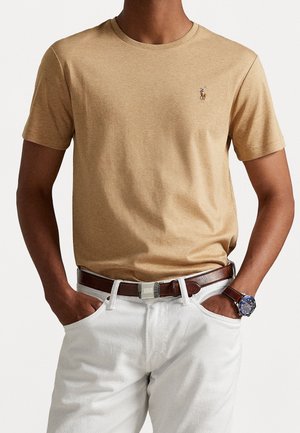 Personne portant un T-shirt à manches courtes beige avec un logo brodé, un pantalon blanc, une ceinture en cuir marron et une montre-bracelet à cadran bleu.