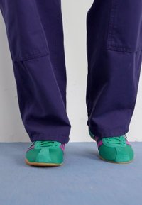 Zeleni nubuk superge s vijoličnimi poudarki in vezalkami, v kombinaciji z ohlapnimi temno vijoličnimi hlačami, na svetlo modri površini.