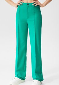 Personne portant un pantalon turquoise taille haute et jambes droites avec un bouton noir et des baskets blanches, mains sur les hanches devant un fond uni.