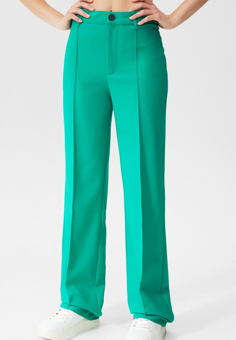 Personne portant un pantalon turquoise taille haute et jambes droites avec un bouton noir et des baskets blanches, mains sur les hanches devant un fond uni.