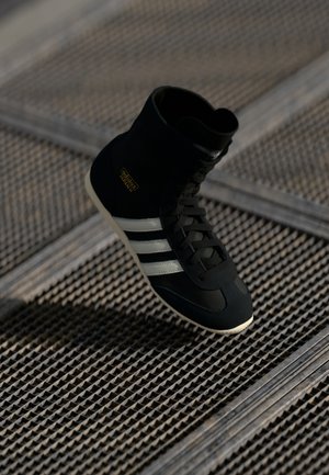 Sort Adidas højtop-sneaker med hvide striber og gul "Adidas Japan" tekst, vist over en struktureret metaloverflade.