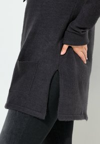 Primo piano di una persona che indossa un maglione grigio scuro a maglia con una fessura laterale e tasca, con una mano poggiata sui fianchi sopra dei pantaloni neri.