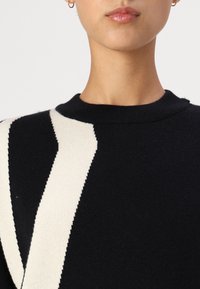 Black sweater with a round neck, featuring a distinct cream zigzag pattern; soft knit texture with fitted cuffs.