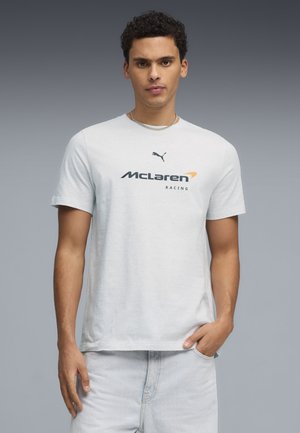 Man draagt lichtgrijze McLaren Racing T-shirt en lichte spijkerbroek, staand met één hand in de zak tegen een effen grijze achtergrond.