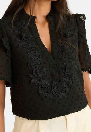 Sort semi-gennemsigtig bluse med blomsterblondebroderi, teksturerede prikker, korte ærmer og flæset høj krave, båret med lyse bukser.