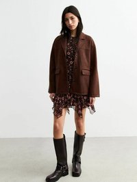 Giacca oversized in camoscio marrone con due tasche frontali, indossata sopra un vestito nero floreale con orlo a frange, abbinata a stivali neri fino al ginocchio.