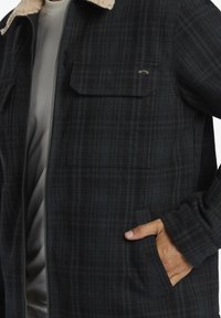 Black and dark green plaid jacket with a cream sherpa collar, zip front, and two chest pockets, featuring a soft texture and button details.