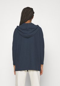 Persona con capelli lunghi e intrecciati, che indossa un maglione con cappuccio blu navy e pantaloni bianchi, mostrata di spalle su uno sfondo semplice.