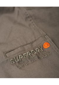 Närbild av grått tyg med sydd ficka och broderat "SUPERDRY" varumärke och logotyp i orange och grå tråd.