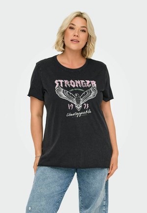 Blond kvinne iført svart T-skjorte med ørne-grafikk og teksten "Stronger than Yesterday 1973 Unstoppable," kombinert med lyseblå jeans.