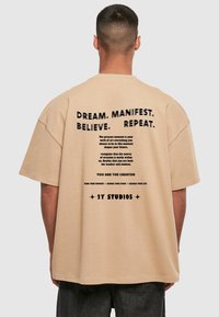 Smėlio spalvos per didelis marškinėlis su juodu tekstu ant nugaros, kuriame yra motyvuojančios frazės ir prekės ženklo 2Y Studios ženklas. Apvalus iškirpimas ir trumpomis rankovėmis.