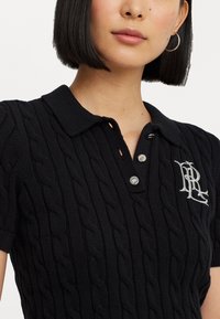 Polo shirt en maille noire avec un motif en câble texturé, patte de boutonnage à trois boutons et logo brodé sur la poitrine gauche. Manches courtes.