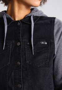 Svart jeansjacka med grå huva inuti, med knappstängning, två framfickor och en dragkedjeficka på bröstet. Strukturerad tyg.