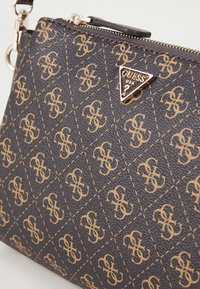 Sac noir texturé avec un motif répétitif de logo doré, fermeture éclair, et un accent en matériel triangulaire doré portant le nom de la marque.