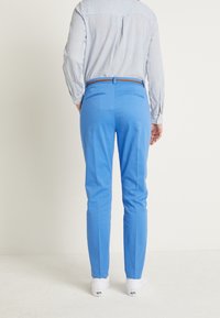 Pantalon tailleur bleu en tissu lisse, coupe slim, avec deux poches arrière et une ceinture marron. Porté avec une chemise rayée.