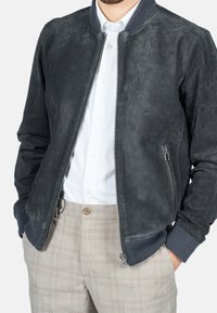 Dunkelgraue Wildleder-Bomberjacke mit gerippten Bündchen und Kragen, ausgestattet mit einem zentralen Reißverschluss und Seitentaschen. Getragen über einem weißen Hemd und hell karierten Hosen.