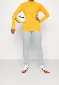 Camiseta amarilla de manga larga de Nike con media cremallera, pantalones ajustados grises, sosteniendo un balón de fútbol texturizado; los zapatos tienen acentos rojos y verdes.