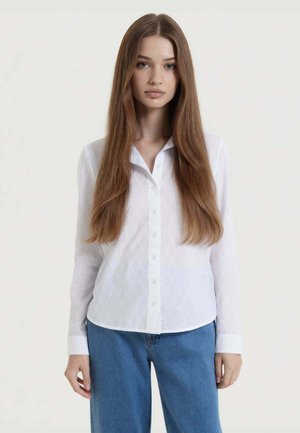 Giovane donna con lunghi capelli castani lisci che indossa una camicia bianca a bottoni con fantasia e jeans blu su uno sfondo semplice.