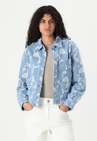 Denim jakna v svetlo modri barvi z belimi cvetličnimi vezeninami, gumbnim zapiranjem in žepom na prsih. Oblečena čez sivo majico in bele hlače.
