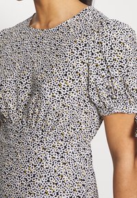 Kukallallinen mekko, jossa on musta, valkoinen ja keltainen kuvio, lyhyet puffihihat ja kerätty yksityiskohta vyötäröllä, valmistettu kevyestä kankaasta.