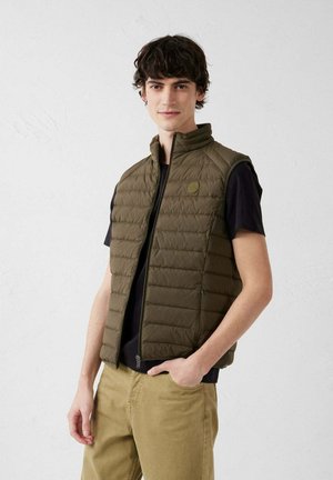 Giovane uomo con capelli scuri che indossa un gilet trapuntato senza maniche verde oliva sopra una t-shirt nera e pantaloni beige, in piedi contro un muro bianco semplice.