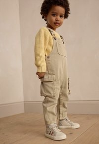 Salopette beige avec de grandes poches, pull en maille texturée jaune, et baskets gris clair avec des graphismes colorés, sur fond neutre.