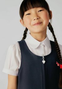Rochie bleumarin cu o cămașă albă cu guler dedesubt, având un charm argintiu în formă de inimă și un accent de gingham roșu. Coafură: împletită.