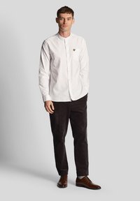 Camicia bianca con colletto alla coreana e tasca con logo giallo; abbinata a pantaloni in velluto a coste scuri e scarpe in pelle marroni.