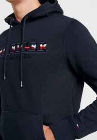 Sudadera azul marino de tela suave, con un logotipo bordado en rojo, blanco y azul, con cordones ajustables y bolsillo frontal.