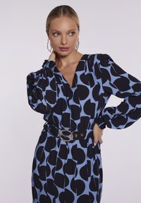 Długa sukienka z długim rękawem w jasnoniebieskim kolorze z czarnymi abstrakcyjnymi kropkami, z głębokim dekoltem w kształcie litery V oraz paskiem w talii z okrągłą złotą klamrą.