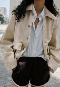 Veste en toile beige avec des pressions, chemise blanche à boutons, et short noir avec une finition blanche. La tenue est décontractée et superposée.