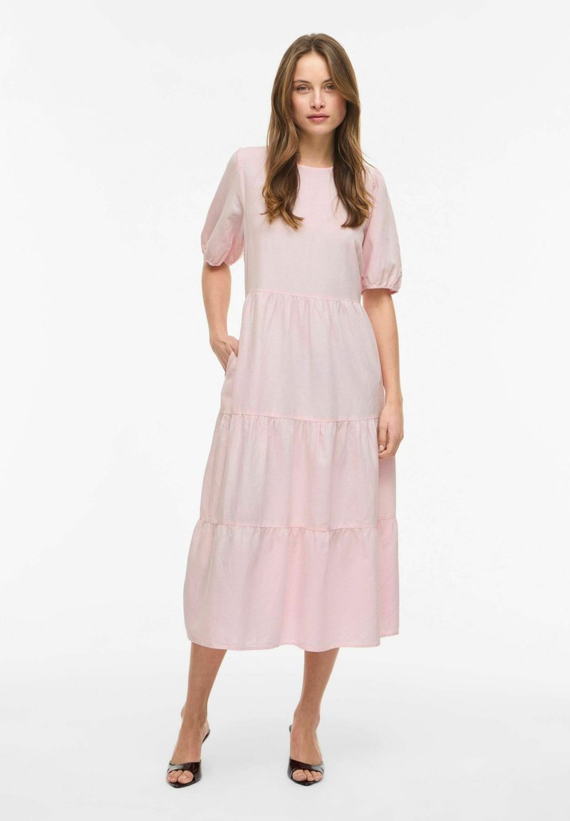 Jeune femme debout portant une robe midi à volants rose clair avec des manches bouffantes et des sandales à talons ouvertes noires sur fond blanc.