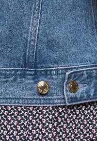 Ourlet de veste en jean avec deux boutons-pression dorés ; texture de tissu bleu avec une jupe à motifs floraux contrastants en bleu marine et blanc avec des accents rouges en dessous.