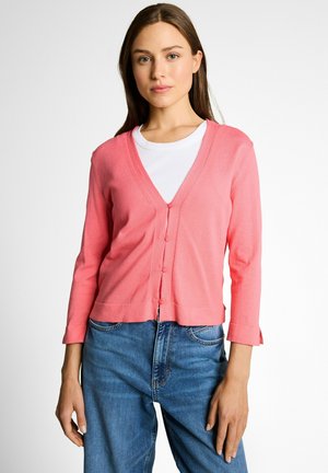 Joven mujer que lleva un cárdigan rosa abotonado sobre una camiseta blanca y vaqueros azules, de pie contra un fondo claro y liso.