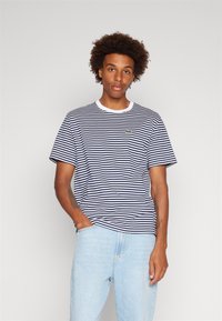 T-shirt rayé bleu marine et blanc avec un col rond blanc et un petit logo vert sur la poitrine gauche. Tissu doux et décontracté ; coupe ample.