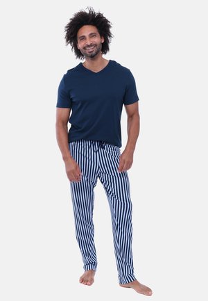 Man staat op blote voeten, draagt een marineblauw shirt met korte mouwen en blauw-witte verticale gestreepte broek, glimlacht tegen een lichte achtergrond.