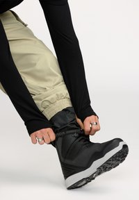 Zwarte waterdichte winterlaars met een textuur bovenkant en een grijze rubberen zool. De persoon past de opgerolde kaki broek aan terwijl hij de laars bevestigt.