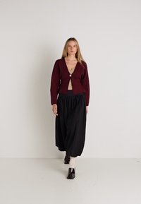 Burgundská pletená cardigan s hlbokým V-výstrihom a jedným gombíkom, v kombinácii s voľnou čiernou sukňou a hnedými topánkami na blokovom podpätku.
