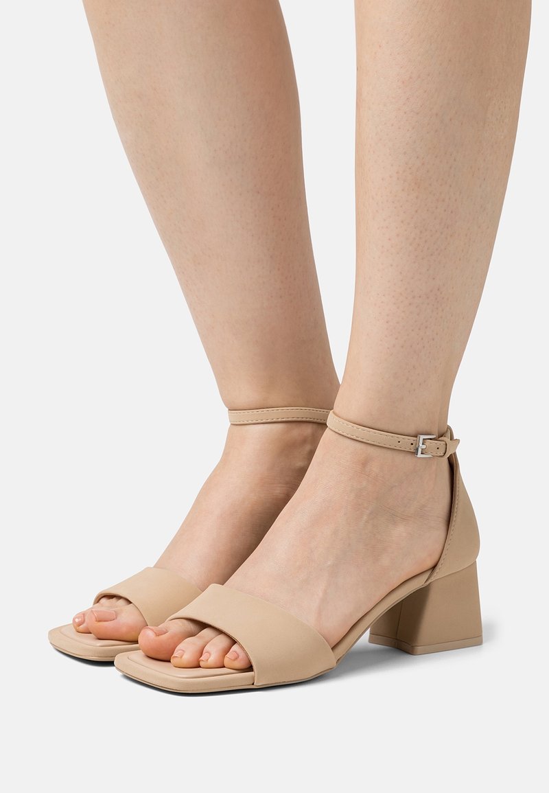 Sandali con tacco aperti beige con una larga fascia sulle dita dei piedi e una cinghia alla caviglia. Le gambe del modello sono parzialmente visibili, mostrando le calzature.