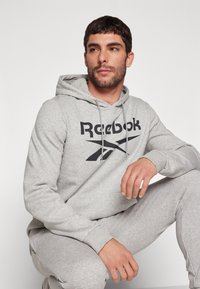 Pilkas džemperis su juodu Reebok logotipu, turintis raištelį ant kepurės ir raukšlėtą rankovių dalį. Suderintas su atitinkamais pilkais treniruočių kelnėmis, siekiant harmoningos išvaizdos.