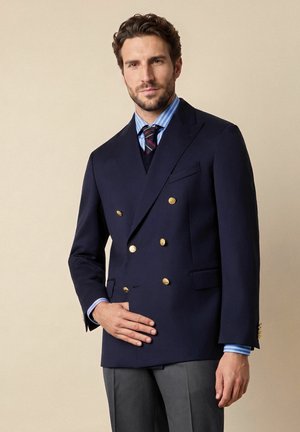 Mann trägt einen marineblauen zweireihigen Blazer mit goldenen Knöpfen, blau gestreiftes Hemd und gemusterte Krawatte, steht vor einem schlichten beigen Hintergrund.