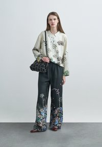 Chemise en soie à imprimé floral avec un col, associée à un pantalon large noir orné d'accents floraux colorés. Le mannequin tient un sac matelassé.