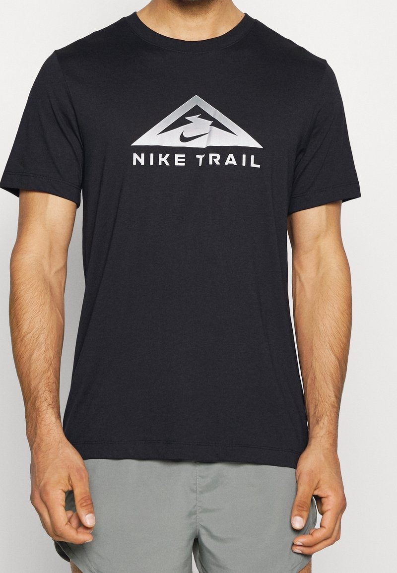 Mand iført en sort Nike Trail t-shirt og grå shorts, stående med armene afslappet langs siderne mod en ensfarvet baggrund.