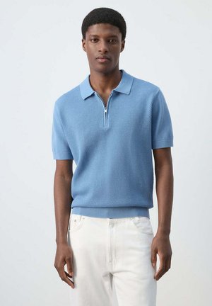 Joven con un jersey tipo polo de manga corta texturizado de color azul claro con cremallera, combinado con pantalones blancos, de pie frente a un fondo liso.