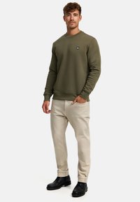 Olivgrüner Sweatshirt mit Rundhals und kleinem Logo, kombiniert mit hellbeigen Hosen und schwarzen Schnürstiefeln. Lässiger Schnitt und Textur.
