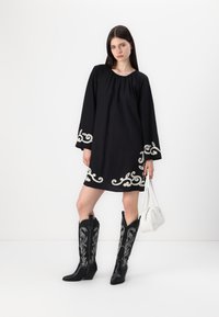 Μαύρο φόρεμα με λευκή λεπτομερή κεντητική περιμετρικά του ποδιού, μακριά μανίκια, συνδυασμένο με μαύρες καουμπόικες μπότες με εκλεπτυσμένη ασημένια ραφή και λευκή τσάντα.